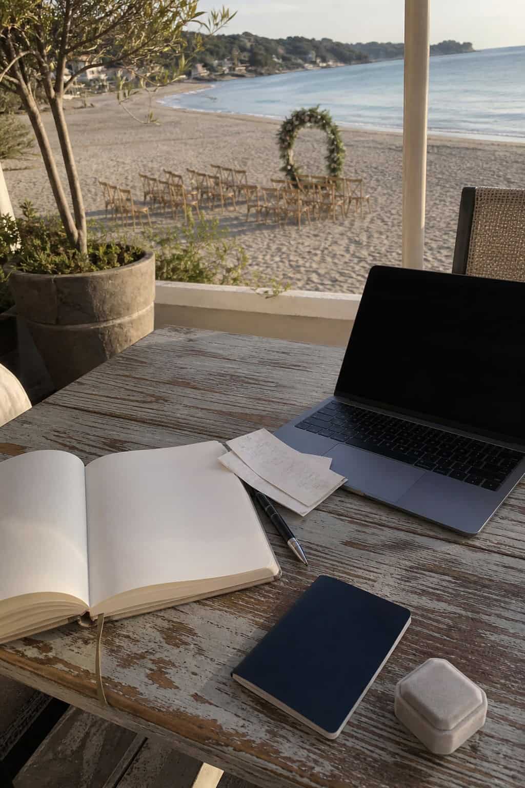 Destination wedding budget notebook, passport, laptop, and ring box overlooking a small beach ceremony