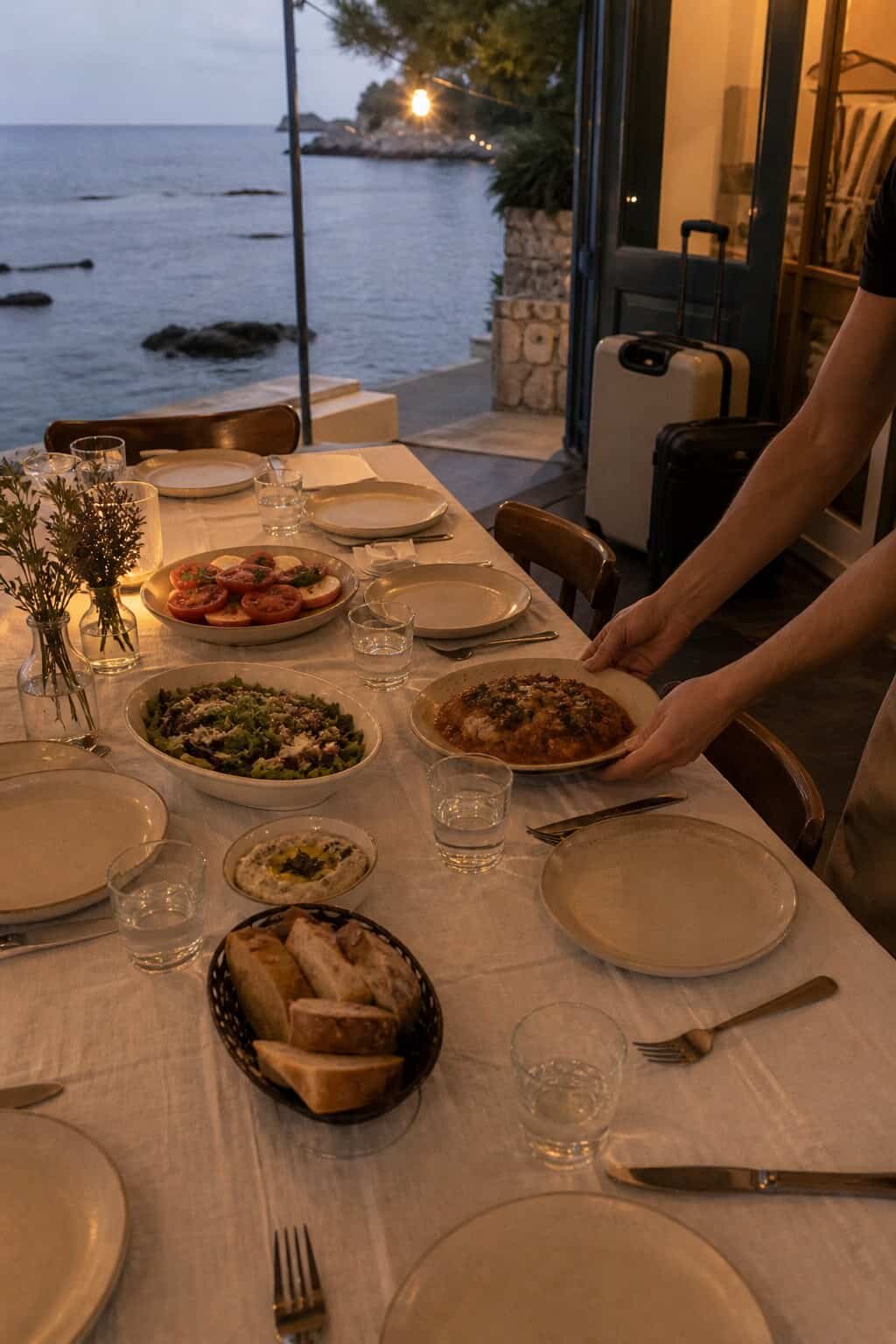 Small welcome dinner being set on a seaside terrace with guests' carry-on luggage nearby