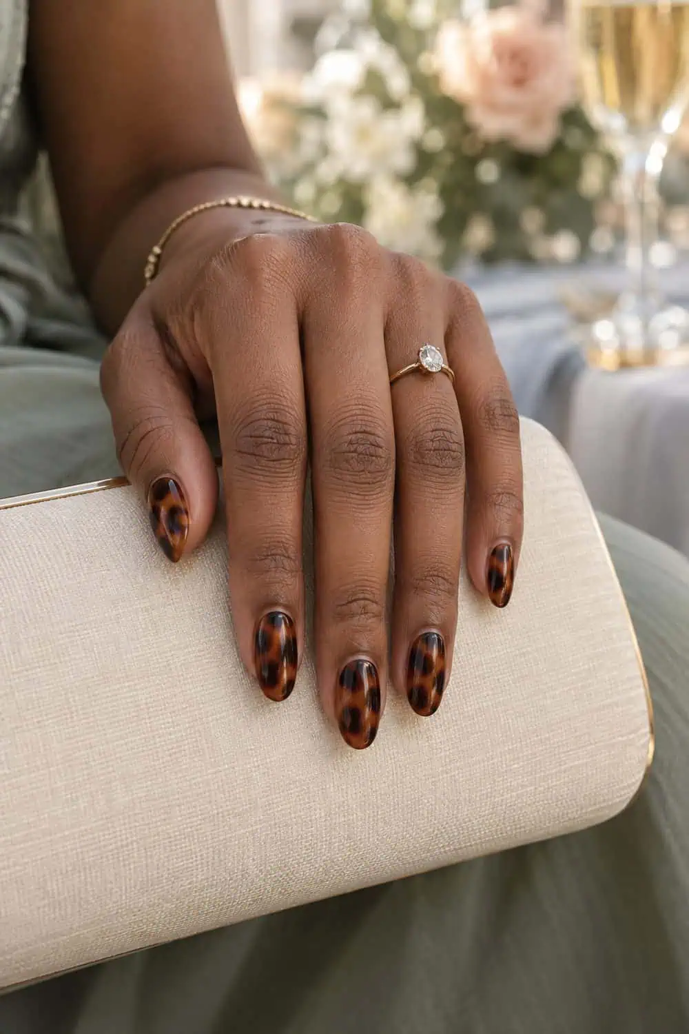 Wedding attendee medium-deep brown hand with medium oval nails in translucent amber-and-brown tortoiseshell pattern, cream clutch