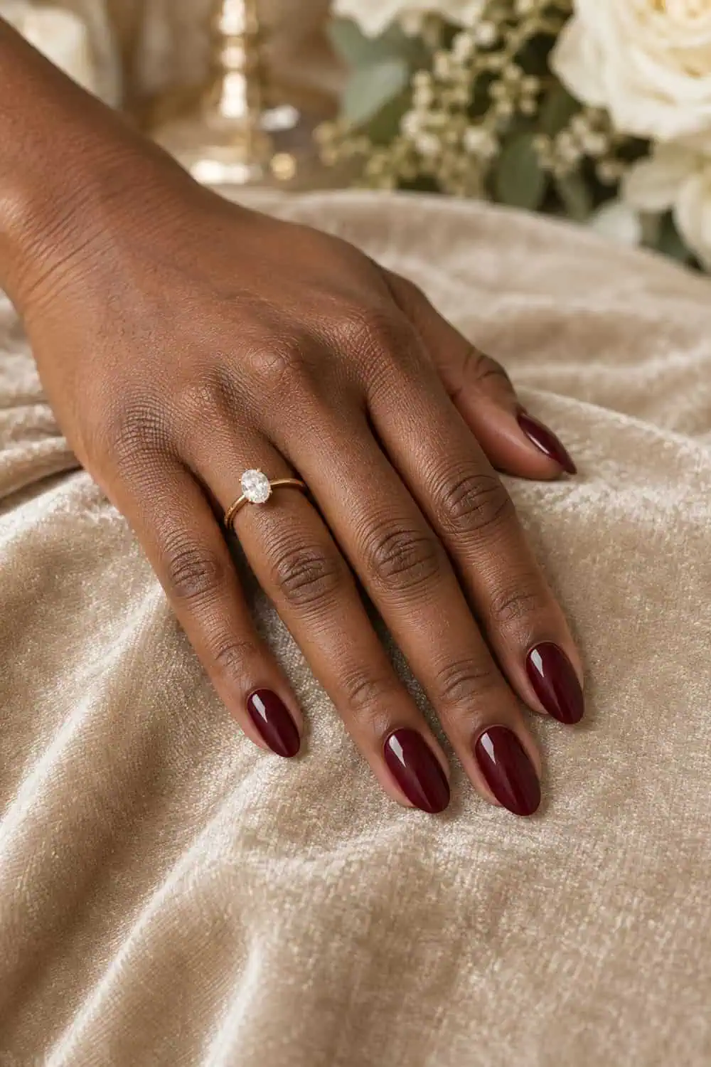 Wedding attendee deep-brown hand with medium almond nails painted solid glossy burgundy, resting on champagne velvet