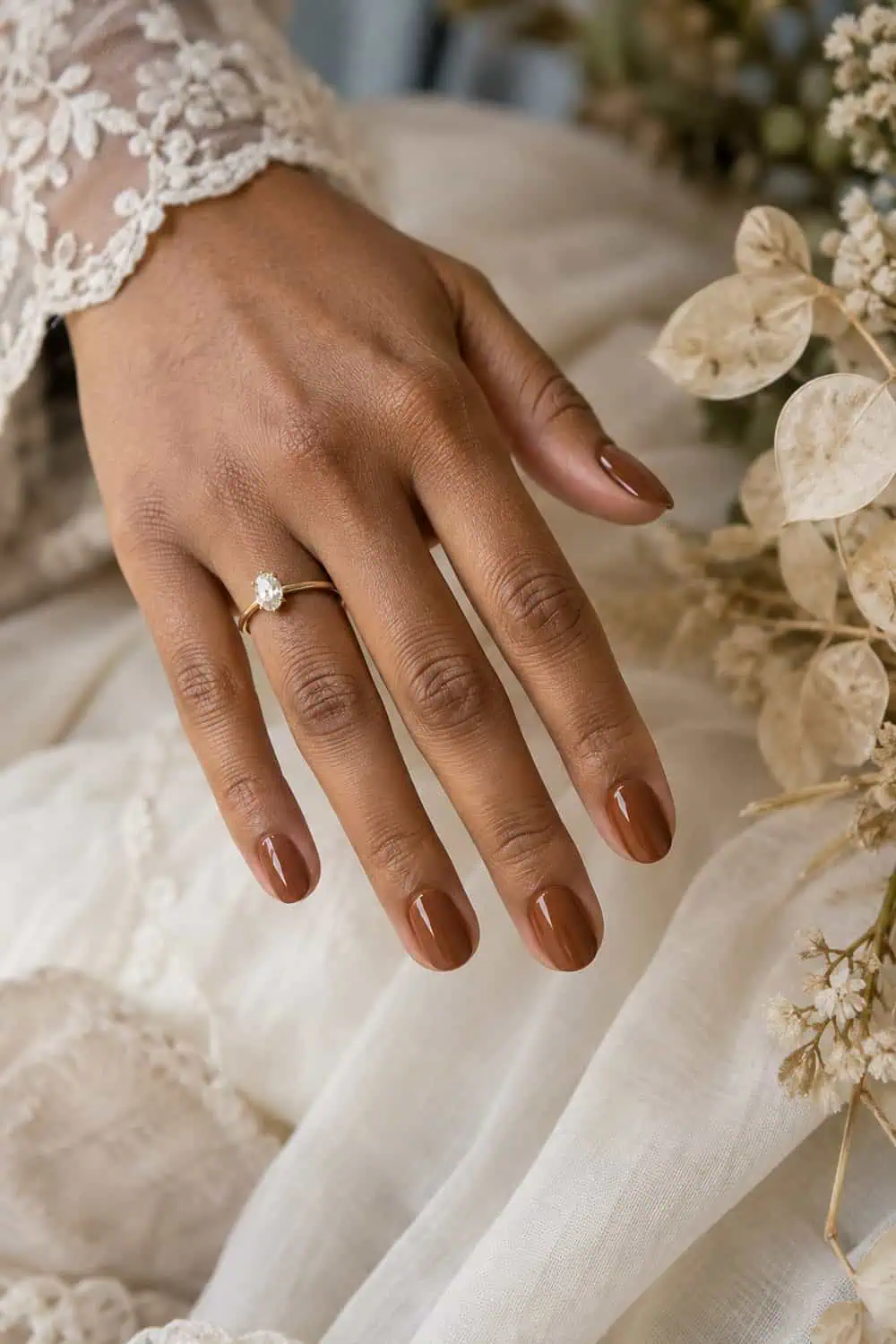 Wedding attendee medium-brown hand with short oval nails painted solid cinnamon brown, dried beige leaves nearby