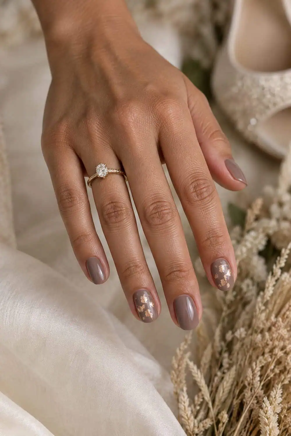 Wedding attendee light-brown hand with short oval nails painted taupe, two accent nails with sparse flat copper foil, dried grasses