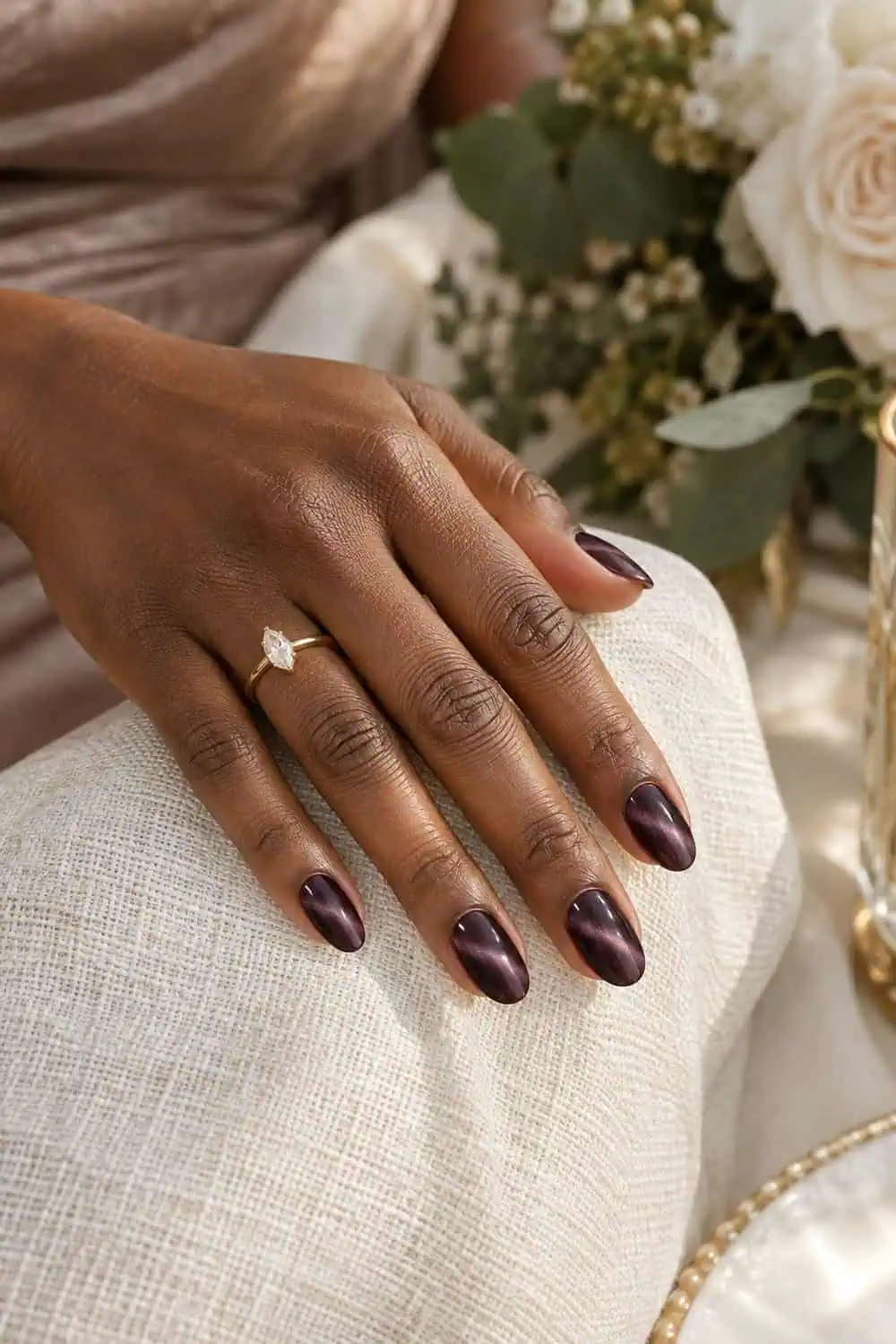 Wedding attendee dark-brown hand with short almond nails painted deep plum magnetic velvet polish, soft light bands visible, cream linen