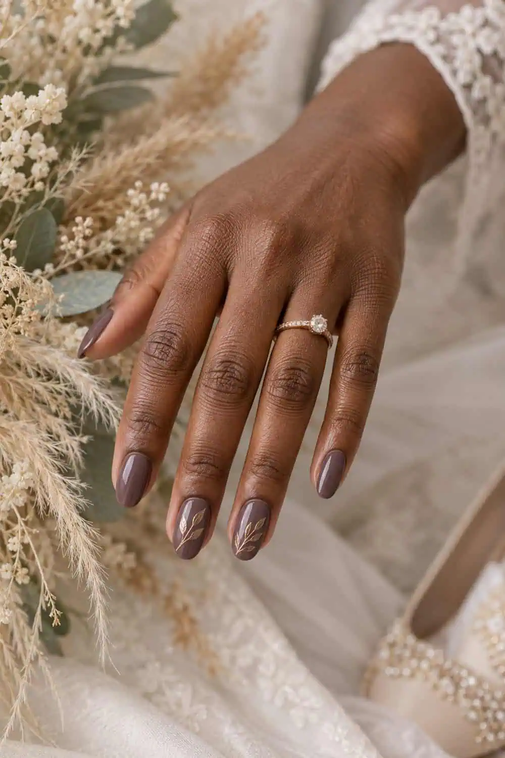 Wedding attendee dark-brown hand with medium oval nails painted smoky mauve, fine bronze leaf-line art on two accents, beige dried grasses
