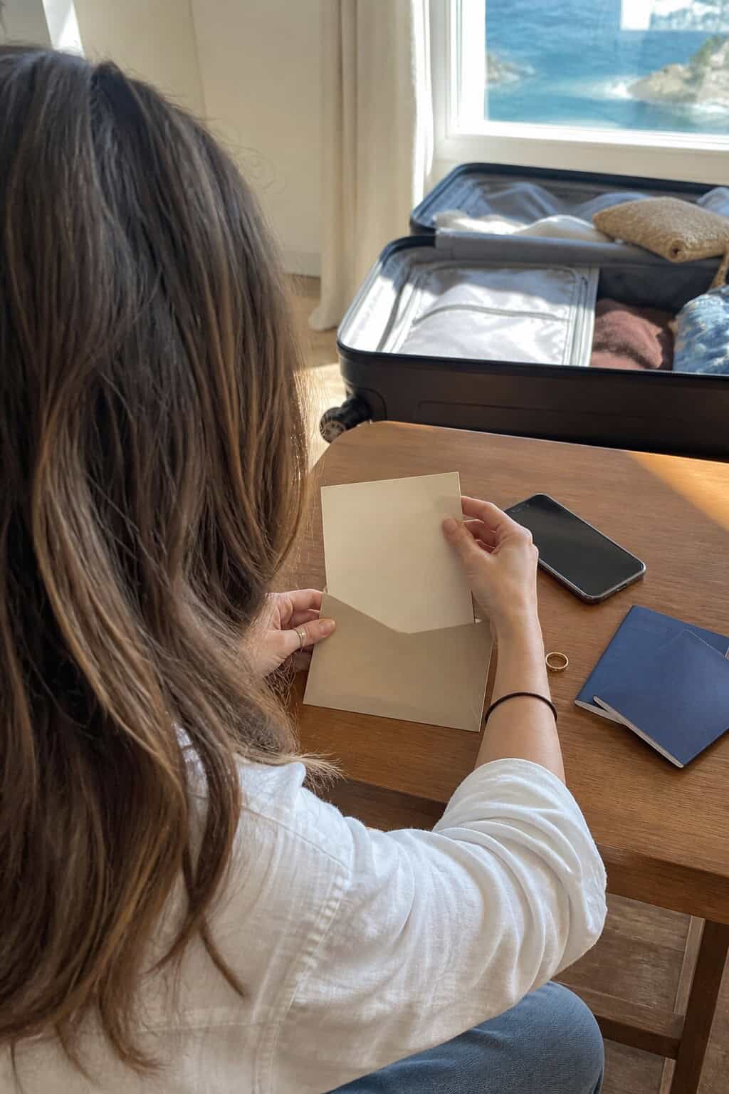 Person placing a destination wedding save-the-date card into an envelope beside passports and a suitcase