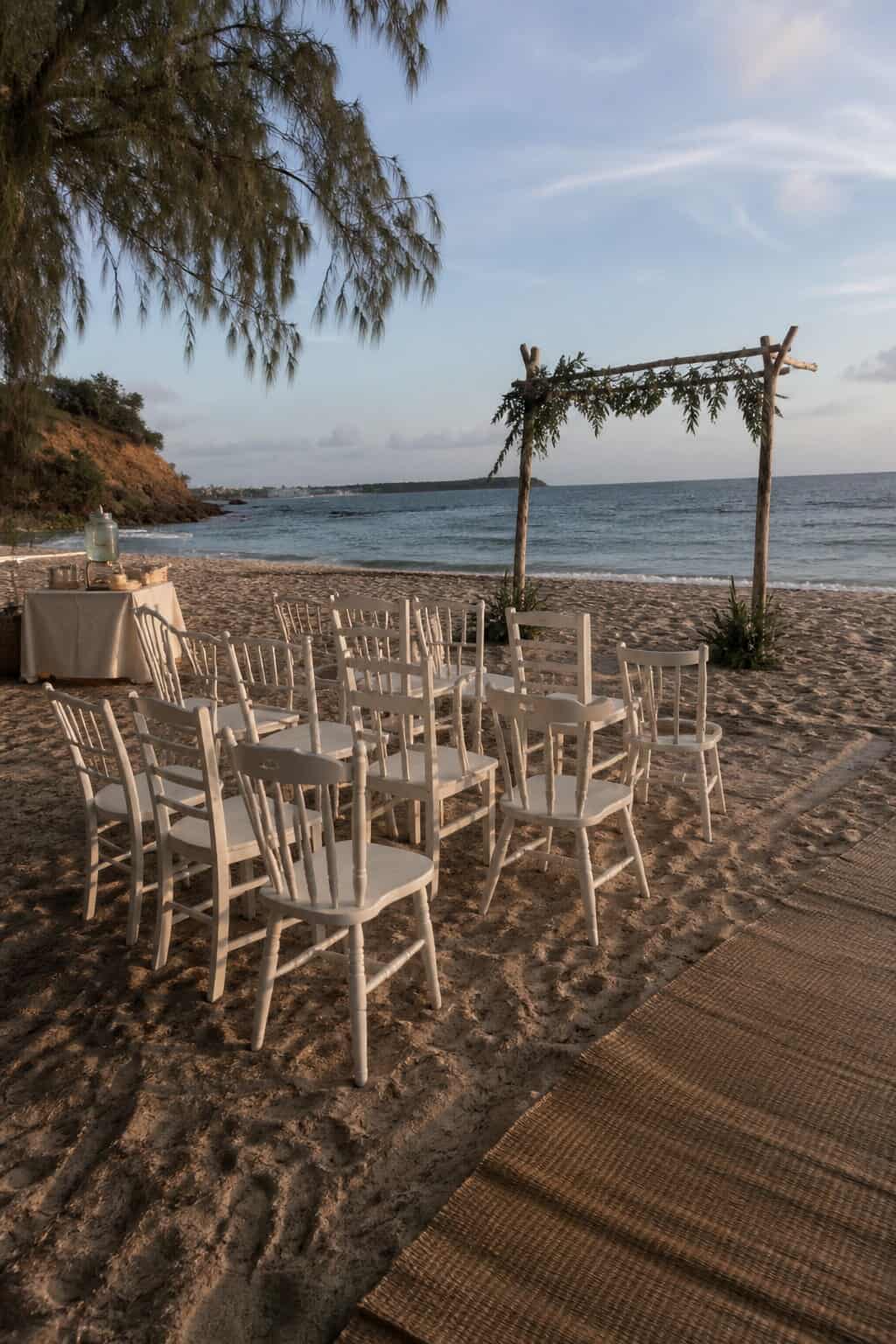 Small beach wedding setup with twelve white chairs, a simple driftwood arch, and an access mat