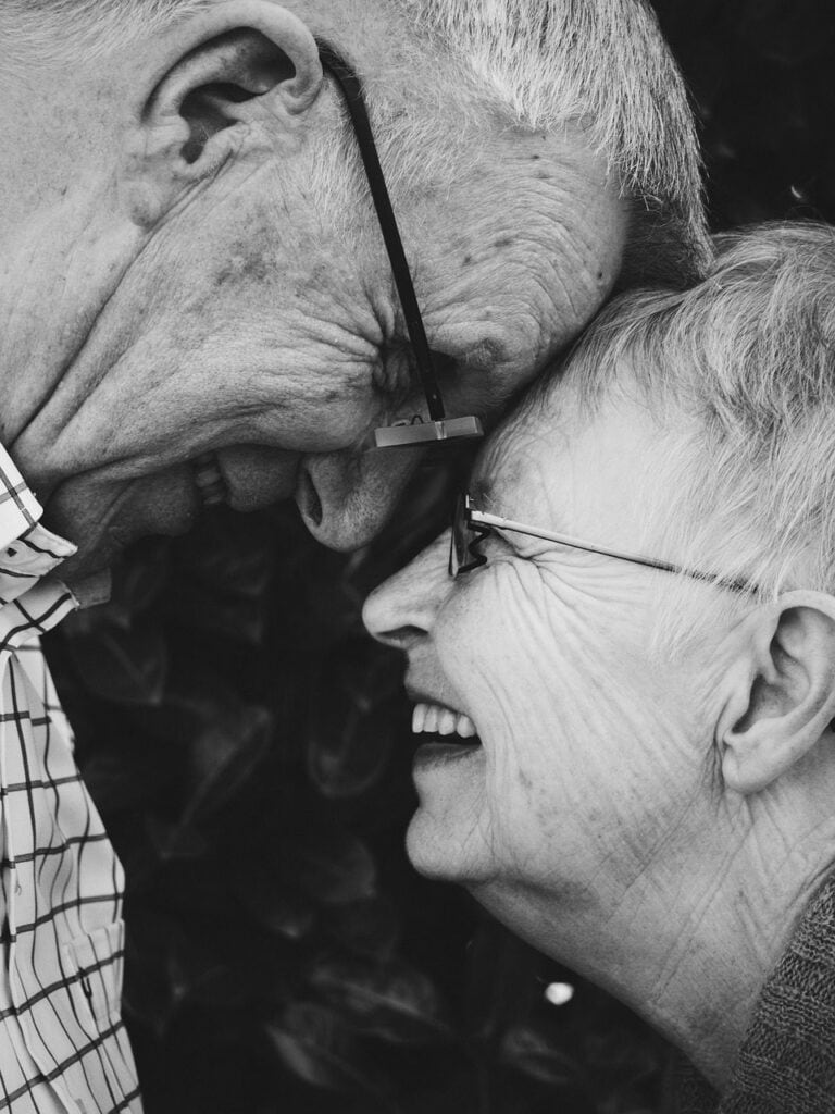 Black and white photo of an elderly couple touching foreheads and laughing together, both wearing glasses